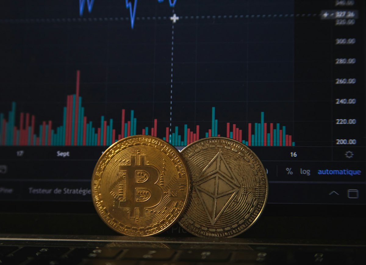 Koin fisik emas Bitcoin dan perak Ethereum di atas keyboard laptop dengan grafik trading pasar keuangan di layar latar belakang